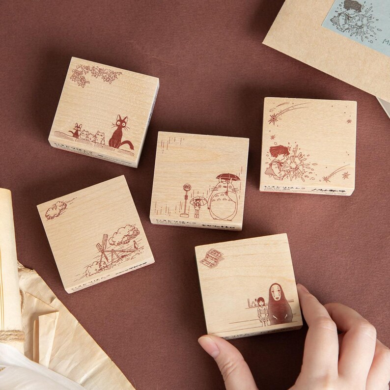 May include: Four square wooden rubber stamps featuring detailed brown ink illustrations of animated characters. The designs include a black cat, a large creature with an umbrella at a bus stop, a person with a star, and a hooded figure with a small companion.