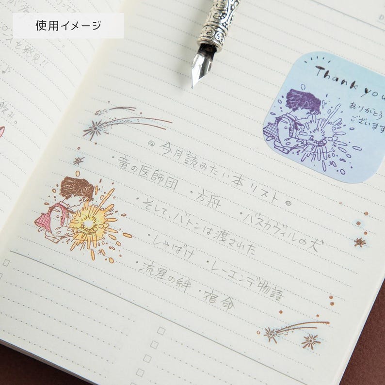 May include: An open notebook with lined pages featuring handwritten Japanese text and colorful illustrations of a person holding a glowing light. A silver fountain pen rests at the top. A blue sticker with the text Thank you is visible.