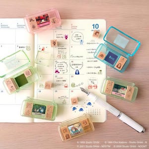Puede incluir: Una colecci&oacute;n de estuches de sellos de pl&aacute;stico transparente en varios colores, cada uno con m&uacute;ltiples sellos con ilustraciones de dibujos animados. Los sellos est&aacute;n dispuestos en un calendario abierto con notas manuscritas y una pluma estilogr&aacute;fica blanca.