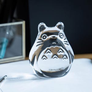 May include: A clear glass figurine of Totoro from Studio Ghibli. The figurine has a rounded body, large eyes, and distinctive markings. The Studio Ghibli logo is visible on the base. The figurine is sitting on a white surface.