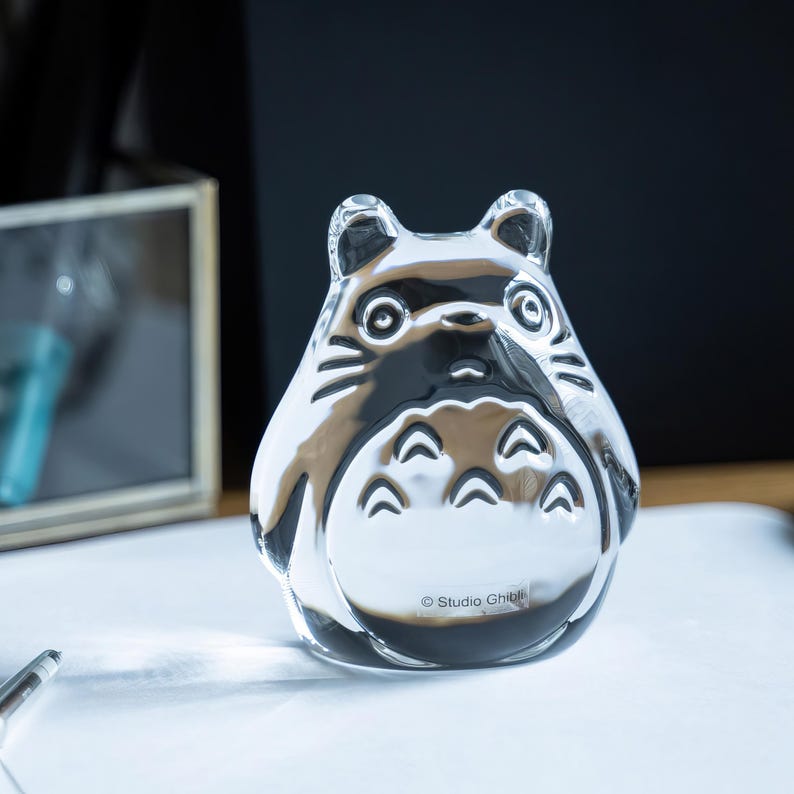 May include: A clear glass figurine of Totoro from Studio Ghibli. The figurine has a rounded body, large eyes, and distinctive markings. The Studio Ghibli logo is visible on the base. The figurine is sitting on a white surface.