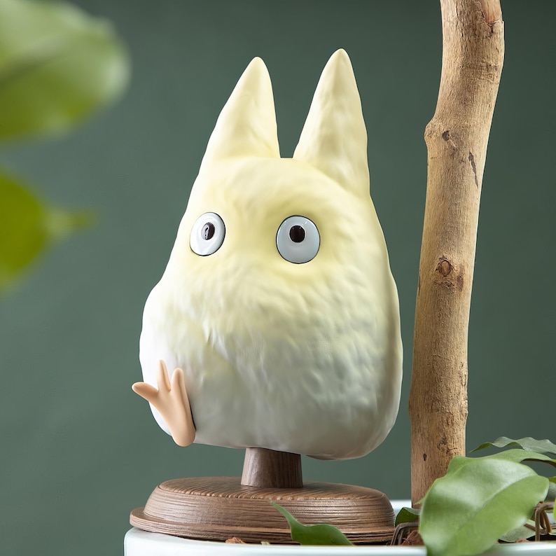 May include: A light yellow figurine of a cartoon character with large eyes and pointed ears. The figurine is mounted on a wooden base and has a small, outstretched hand. The background is a muted green.