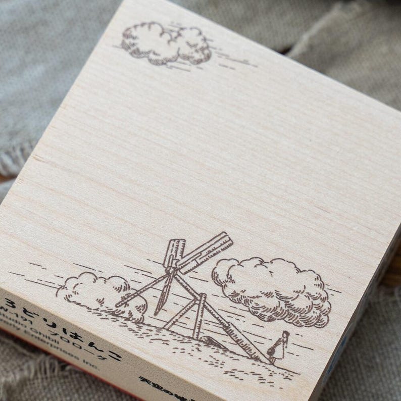 May include: A square wooden rubber stamp featuring a detailed line art illustration of a windmill, clouds, and a small figure. The stamp includes Japanese text and the Studio Ghibli copyright information printed in black ink on the light wood surface.