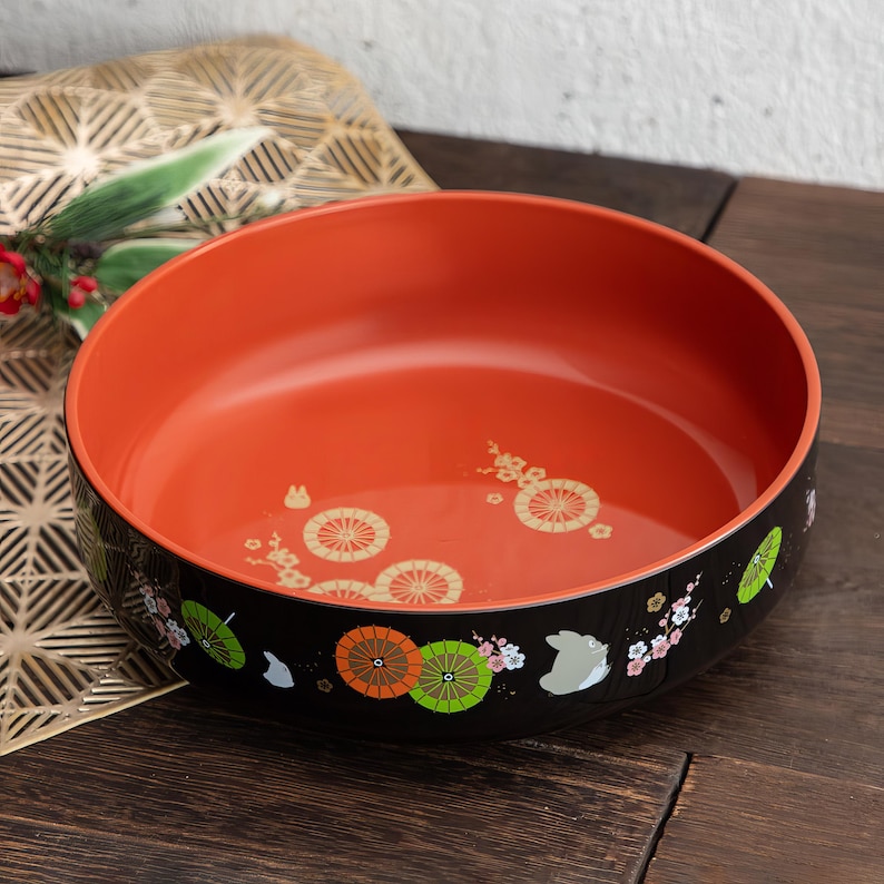 May include: A round, orange and black bowl with a decorative design. The interior is a vibrant orange, while the exterior is black with illustrations of umbrellas, flowers, and a rabbit. The bowl sits on a dark wooden surface.
