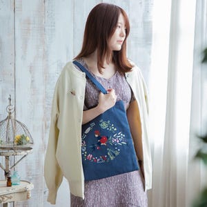 K&ouml;nnte beinhalten: Eine Frau tr&auml;gt einen wei&szlig;en Cardigan und ein Blumenkleid und h&auml;lt eine blaue Tragetasche mit einem gestickten Design eines M&auml;dchens, das auf einem Besen mit Blumen und anderen fantasievollen Elementen fliegt.