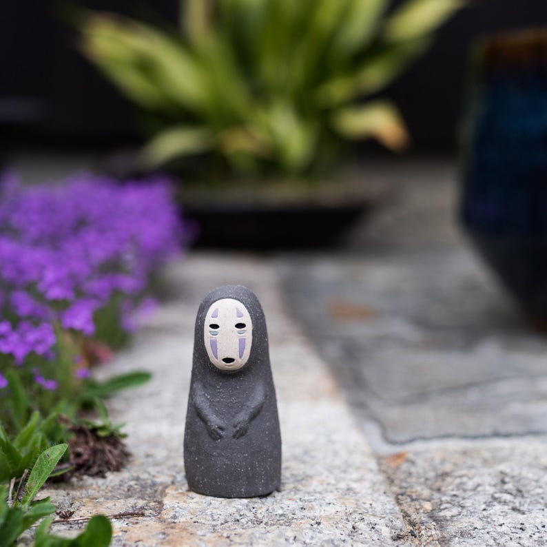 Puede incluir: Una peque&ntilde;a figura de color gris oscuro con una superficie texturizada, que presenta un rostro p&aacute;lido de forma ovalada con dos marcas verticales moradas bajo los ojos y una peque&ntilde;a boca negra. La figura tiene una parte superior redondeada.