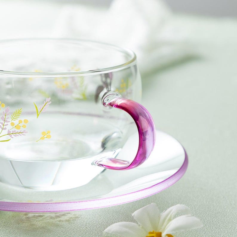 Op de afbeelding: Een theekop van helder glas met een fel magenta handvat en een bijpassende schotel met een lichtroze rand. De kop is versierd met een delicaat botanisch patroon van kleine gele bloemetjes en zachtpaarse takjes op de buitenkant.