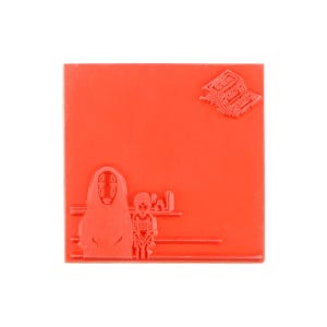 May include: A square red rubber stamp featuring a raised design of a masked figure standing next to a smaller character. In the upper right corner, there is a small, detailed graphic of a building structure with Japanese text characters.