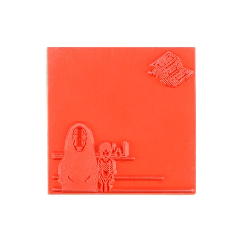 May include: A square red rubber stamp featuring a raised design of a masked figure standing next to a smaller character. In the upper right corner, there is a small, detailed graphic of a building structure with Japanese text characters.