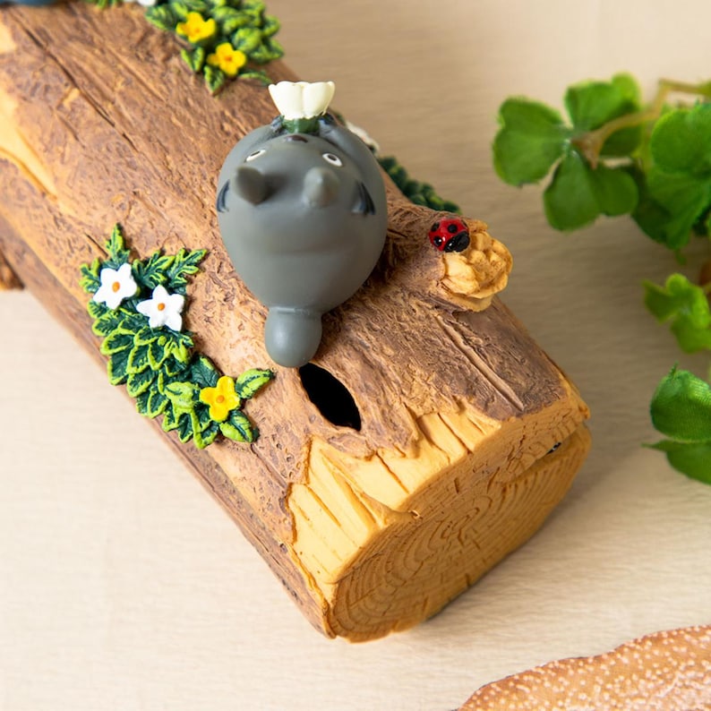 May include: A gray Totoro figure rests on a decorative log, adorned with green foliage, white and yellow flowers, and a small ladybug. The log has a textured, wood-grain appearance and a small hole. A whimsical, nature-themed collectible.