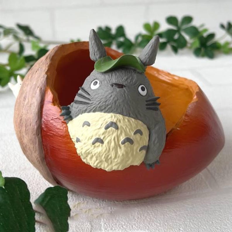K&ouml;nnte beinhalten: Graue Totoro-Figur mit gr&uuml;nem Blatthut in einem dekorativen roten und orangefarbenen Beh&auml;lter. Der Beh&auml;lter hat einen braunen Rand und eine strukturierte Oberfl&auml;che. Die Figur hat einen cremefarbenen Bauch mit schwarzen Markierungen.