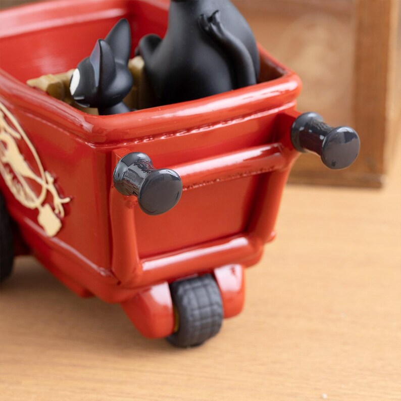 May include: A red decorative wagon with black wheels and handles. Two black cat figurines are inside the wagon. The wagon has a gold emblem on the side. The wagon is a decorative item.