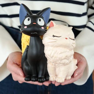 Puede incluir: Dos figuras de gato, una negra con ojos grandes y orejas bordeadas de azul, y otra de color crema con pelo largo y ojos cerrados. El gato negro tiene acentos rosados en las mejillas y una nariz peque&ntilde;a, mientras que el gato crema tiene bigotes finos.