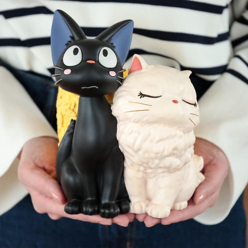 Puede incluir: Dos figuras de gato, una negra con ojos grandes y orejas bordeadas de azul, y otra de color crema con pelo largo y ojos cerrados. El gato negro tiene acentos rosados en las mejillas y una nariz peque&ntilde;a, mientras que el gato crema tiene bigotes finos.