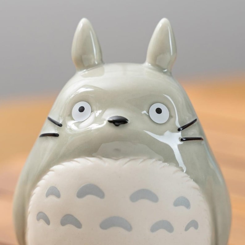 Puede incluir: Figura de cer&aacute;mica de Totoro, personaje de la pel&iacute;cula de animaci&oacute;n Mi Vecino Totoro. La figura es de color gris claro con ojos, nariz y bigotes negros. Tiene dos orejas puntiagudas y una barriga blanca con marcas grises.