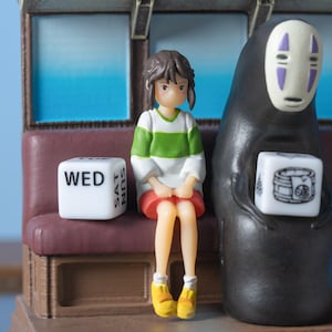 May include: A figurine set featuring characters from a Japanese animated film. A girl in a green and white striped shirt and red skirt sits next to a black figure with a white mask. A white die with the word "WED" is visible.