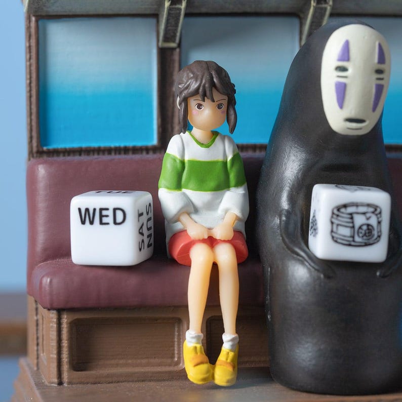 May include: A figurine set featuring characters from a Japanese animated film. A girl in a green and white striped shirt and red skirt sits next to a black figure with a white mask. A white die with the word "WED" is visible.
