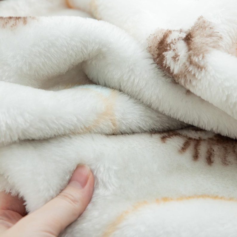 Peut inclure: Gros plan sur une couverture blanche et douce avec une texture en peluche. La couverture pr&eacute;sente un motif subtil dans les tons marron et beige. Le tissu semble chaud et invitant.
