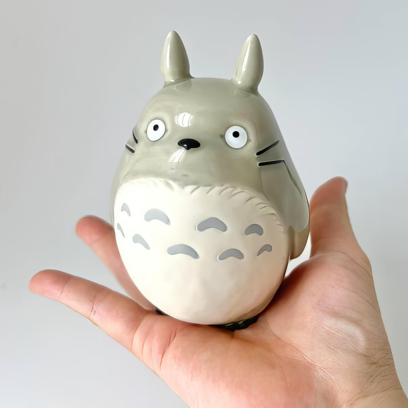 Puede incluir: Figura de Totoro de cer&aacute;mica gris con vientre blanco y marcas grises. La figura tiene ojos grandes y redondos, orejas peque&ntilde;as y bigotes negros. La figura se sostiene en la mano abierta de una persona.
