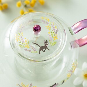 Puede incluir: Una tetera de cristal transparente con tapa y pomo morado, decorada con una ilustraci&oacute;n de un gato negro, flores de mimosa amarillas y ramas de lavanda moradas. La tetera tiene un asa morada transl&uacute;cida y motivos florales a juego.