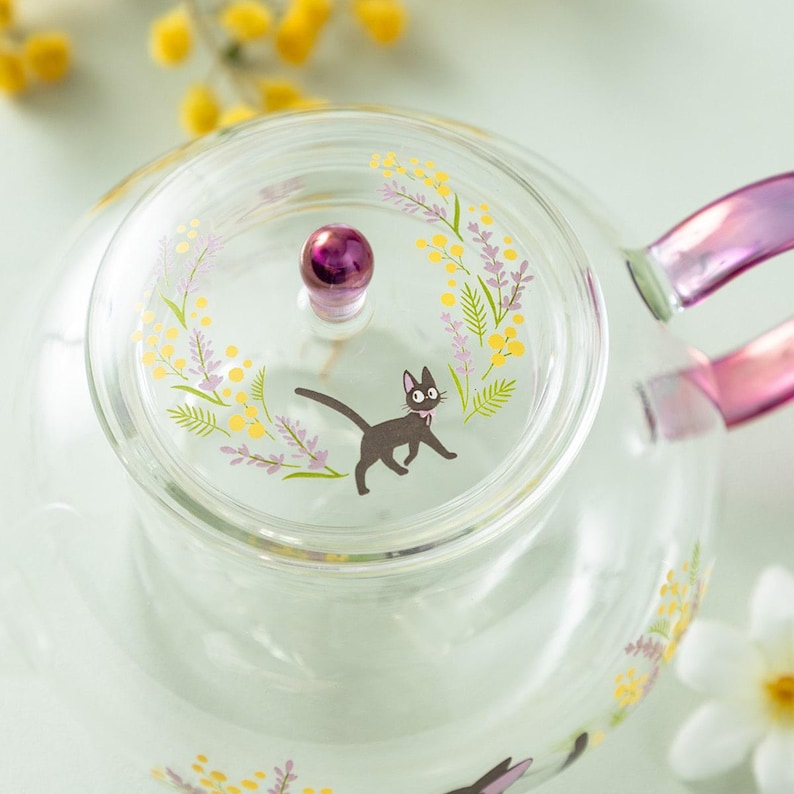 Puede incluir: Una tetera de cristal transparente con tapa y pomo morado, decorada con una ilustraci&oacute;n de un gato negro, flores de mimosa amarillas y ramas de lavanda moradas. La tetera tiene un asa morada transl&uacute;cida y motivos florales a juego.