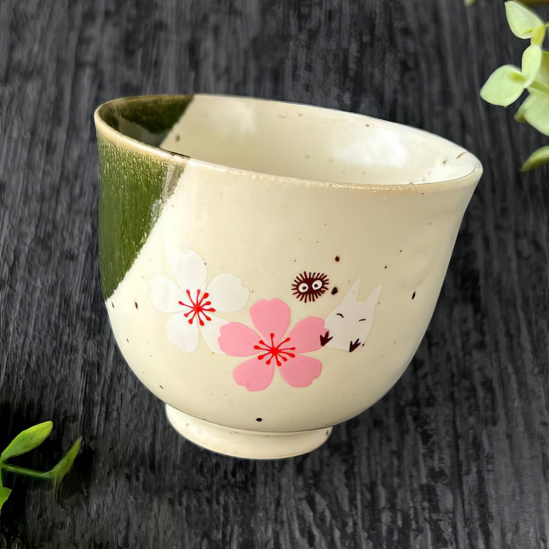 May include: Cream-colored ceramic bowl with a green accent on the rim. The bowl features pink and white floral designs and a cartoon character illustration. The bowl has a small, rounded base.