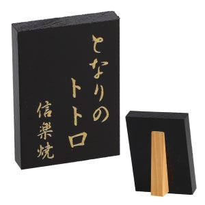 May include: Black wooden sign with gold Japanese characters. The sign is rectangular and has a wooden stand on the back. The text on the sign reads "となりのトトロ" and "信楽焼".