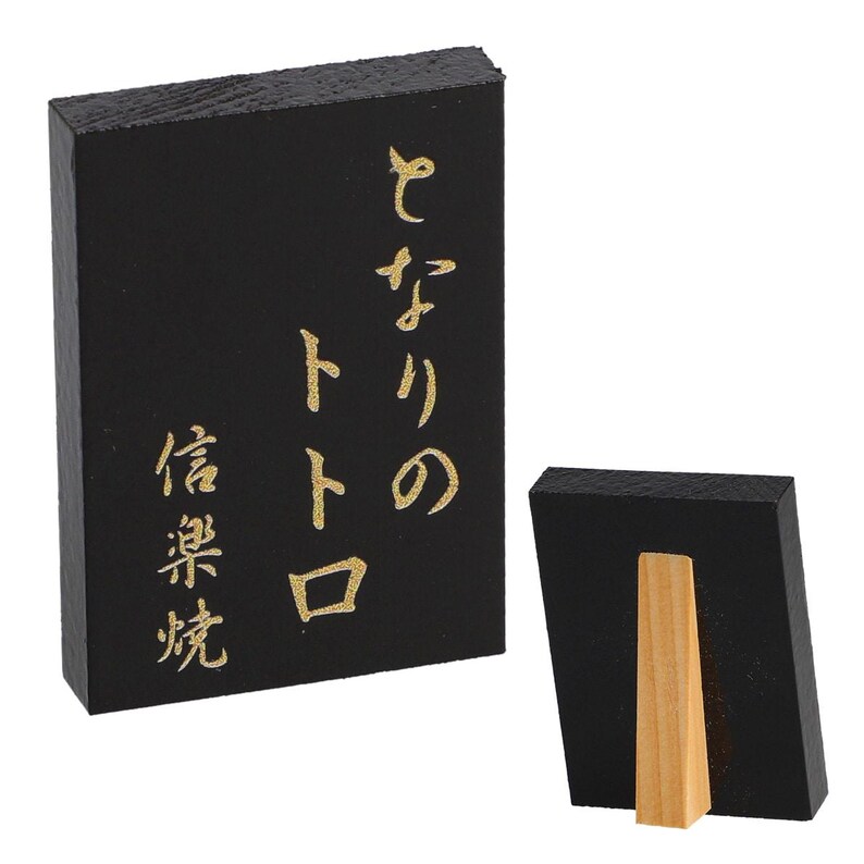May include: Black wooden sign with gold Japanese characters. The sign is rectangular and has a wooden stand on the back. The text on the sign reads "となりのトトロ" and "信楽焼".