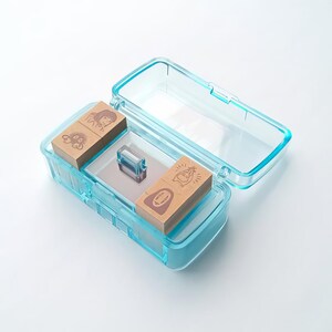 Puede incluir: Una caja de sellos de pl&aacute;stico azul transparente con tapa abatible, que contiene cuatro sellos de madera con ilustraciones de personajes de dibujos animados. La caja tambi&eacute;n contiene una peque&ntilde;a almohadilla de tinta transparente y una superficie espejada.