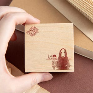 May include: A square wooden rubber stamp featuring a dark brown engraved illustration of a small girl sitting next to a tall, hooded figure. In the top left corner, there is a small rectangular stamp design with Japanese text and a train ticket icon.