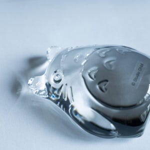 May include: A clear glass figurine in the shape of a cartoon character, with rounded features and embossed details. The figurine has a shiny, reflective surface and is set against a light background. The text "Studio Ghibli" is visible on the bottom.