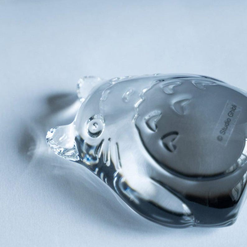 May include: A clear glass figurine in the shape of a cartoon character, with rounded features and embossed details. The figurine has a shiny, reflective surface and is set against a light background. The text "Studio Ghibli" is visible on the bottom.