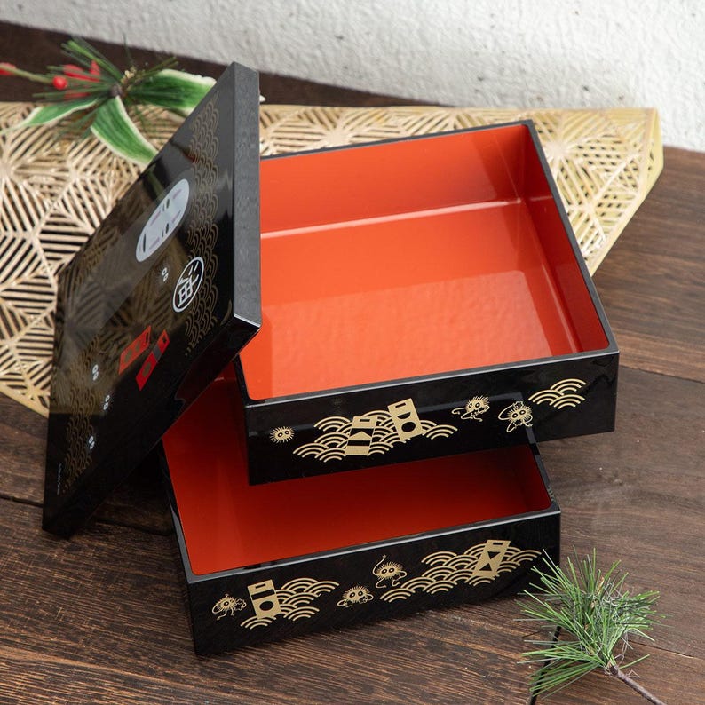 May include: A black lacquered Japanese bento box with a vibrant orange interior. The box features gold wave and stylized design elements. The lid is slightly ajar, revealing the interior. The bento box is on a wooden surface.