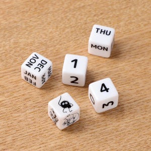 May include: Five white dice with black text and illustrations are arranged on a light brown wooden surface. The dice display numbers, months, and days of the week. One die features a cartoon character.