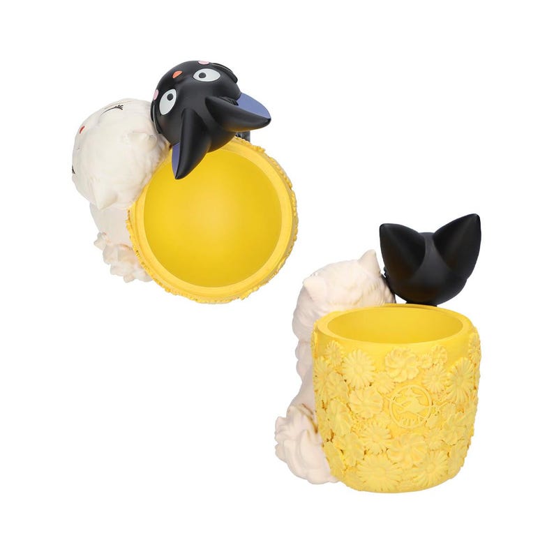Puede incluir: Un recipiente cil&iacute;ndrico amarillo con una superficie floral texturizada y dos figuras de gato unidas al lateral. Un gato es negro con orejas interiores moradas y el otro es blanco. El recipiente tiene un interior liso y un exterior esculpido.