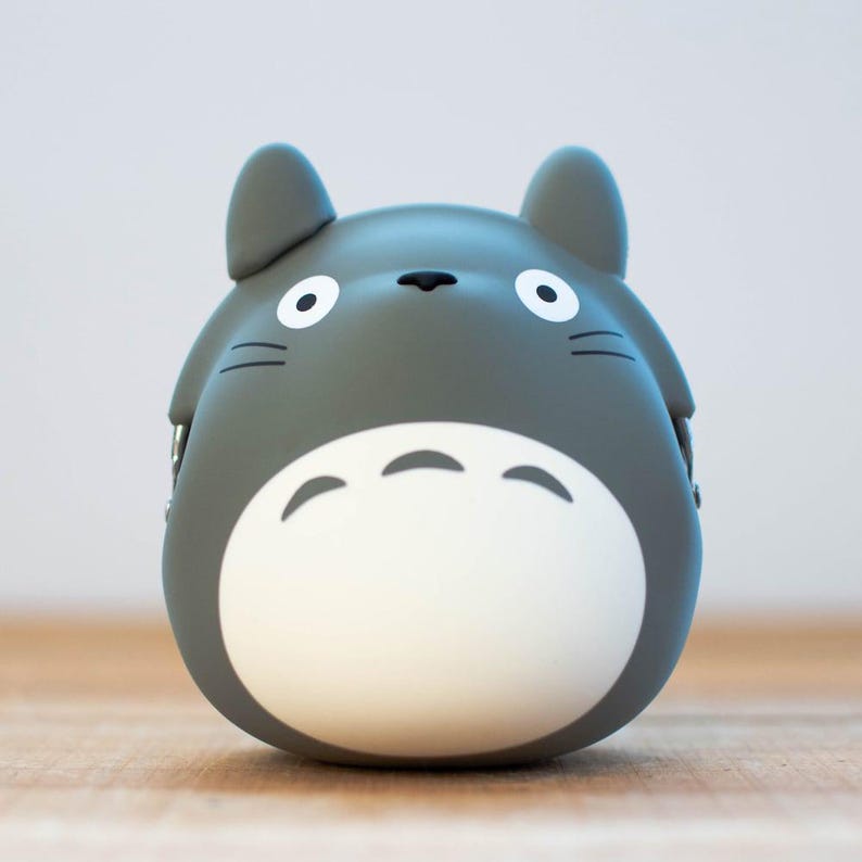 May include: A gray Totoro-shaped coin purse with a white belly and large, round eyes. The purse has small ears and whiskers, and is sitting on a wooden surface. The design is based on the animated film My Neighbor Totoro.