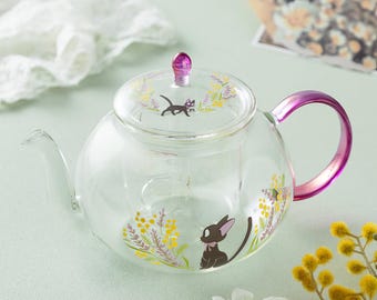 May include: A clear glass teapot featuring a black cat illustration, yellow floral accents, and a metallic purple handle. The lid includes a matching purple knob and a small black cat graphic. The design is printed on the transparent glass body.