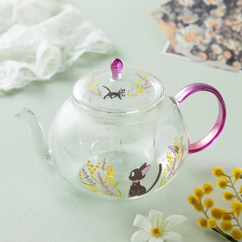 Puede incluir: Una tetera de cristal transparente con una ilustraci&oacute;n de un gato negro, detalles florales amarillos y un asa de color morado met&aacute;lico. La tapa incluye un pomo morado a juego y un peque&ntilde;o gr&aacute;fico de gato negro sobre el cristal.