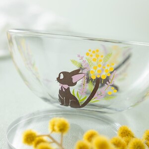May include: A clear glass bowl featuring a printed graphic of a small black cat with a pink bow, surrounded by illustrations of yellow mimosa flowers and light purple lavender sprigs. The bowl has a smooth, curved rim and transparent walls.