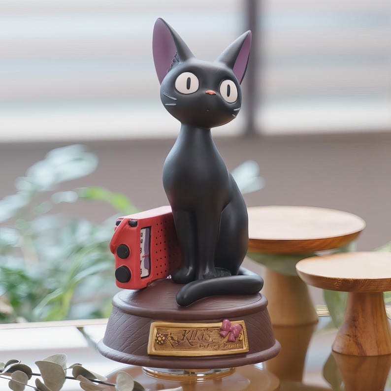 May include: A black cat figurine with large white eyes and pink-lined ears sits atop a brown base. A red box is positioned next to the cat. The base has a gold plaque with text and a pink bow. The figurine is a decorative item.