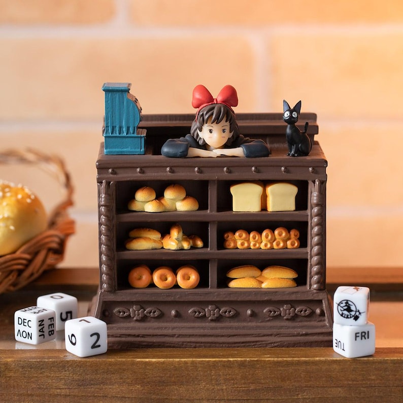 Pode incluir: Uma exibi&ccedil;&atilde;o de padaria em miniatura com uma menina com um la&ccedil;o vermelho e um gato preto. A vitrine marrom est&aacute; cheia de p&atilde;es e donuts. Uma caixa registradora azul est&aacute; no topo. Dados presentes.