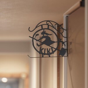 K&ouml;nnte beinhalten: Schwarze Metall-Wanddekoration mit einer Hexe auf einem Besen, darunter das Wort "KIKI". Das Design ist kreisf&ouml;rmig mit dekorativen Elementen. Die Dekoration ist an einem wei&szlig;en T&uuml;rrahmen befestigt.