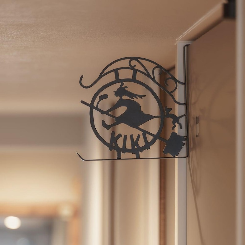 K&ouml;nnte beinhalten: Schwarze Metall-Wanddekoration mit einer Hexe auf einem Besen, darunter das Wort "KIKI". Das Design ist kreisf&ouml;rmig mit dekorativen Elementen. Die Dekoration ist an einem wei&szlig;en T&uuml;rrahmen befestigt.