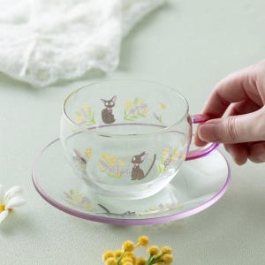 Op de afbeelding: Een theekop en bijpassende schotel van helder glas met een fijne print van kleine zwarte katjes met roze strikjes, omringd door gele en paarse bloemtakjes. Het oortje van de kop en de rand van de schotel hebben een subtiele roze kleurverloop.