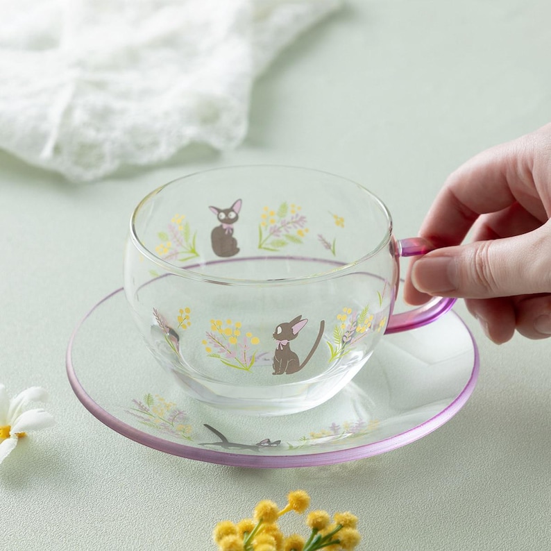 Op de afbeelding: Een theekop en bijpassende schotel van helder glas met een fijne print van kleine zwarte katjes met roze strikjes, omringd door gele en paarse bloemtakjes. Het oortje van de kop en de rand van de schotel hebben een subtiele roze kleurverloop.