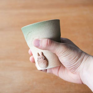 May include: A ceramic cup with a light green top and a beige body. The cup features a raised relief of a cartoon character on the side. The cup is held in a hand.