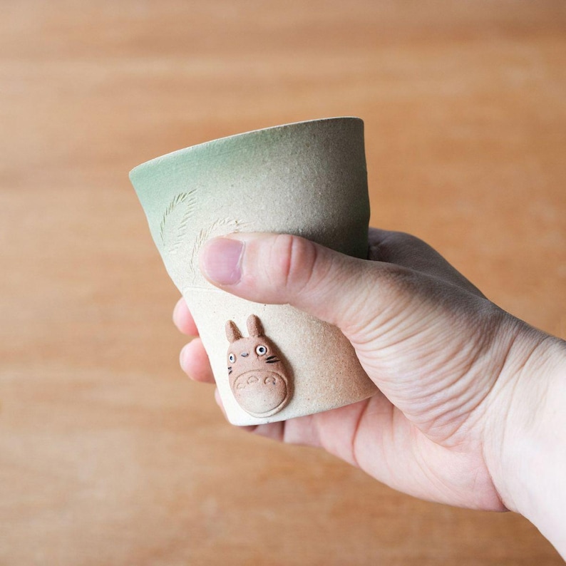 May include: A ceramic cup with a light green top and a beige body. The cup features a raised relief of a cartoon character on the side. The cup is held in a hand.