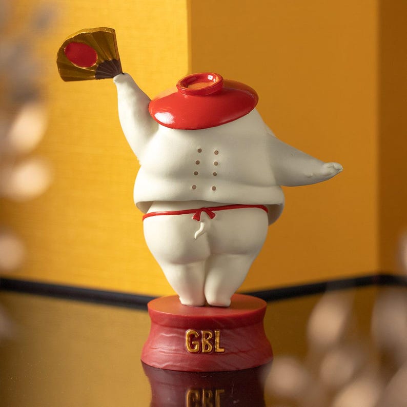 Peut inclure: Figurine blanche repr&eacute;sentant un lutteur de sumo avec un chapeau et un pagne rouges, tenant un &eacute;ventail. La figurine est sur une base rouge avec les lettres "GBL". L'&eacute;ventail est dor&eacute; et rouge.