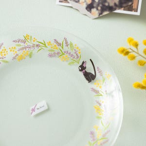 May include: A clear glass plate featuring a printed design of a small black cat with a purple bow, surrounded by a curved arrangement of yellow mimosa flowers and purple lavender sprigs. A small rectangular white label is attached to the glass surface.