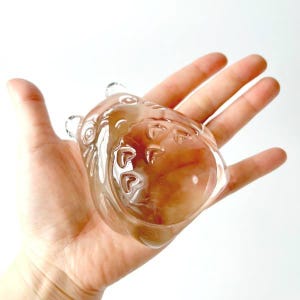 May include: A clear, glass figurine in the shape of a bear, held in a hand. The bear has etched details and a light brown hue. The figurine is a decorative item.